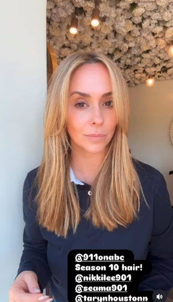 Jennifer Love Hewitt with blonde hair, wearing a dark blue shirt, standing in a room with floral ceiling decor.