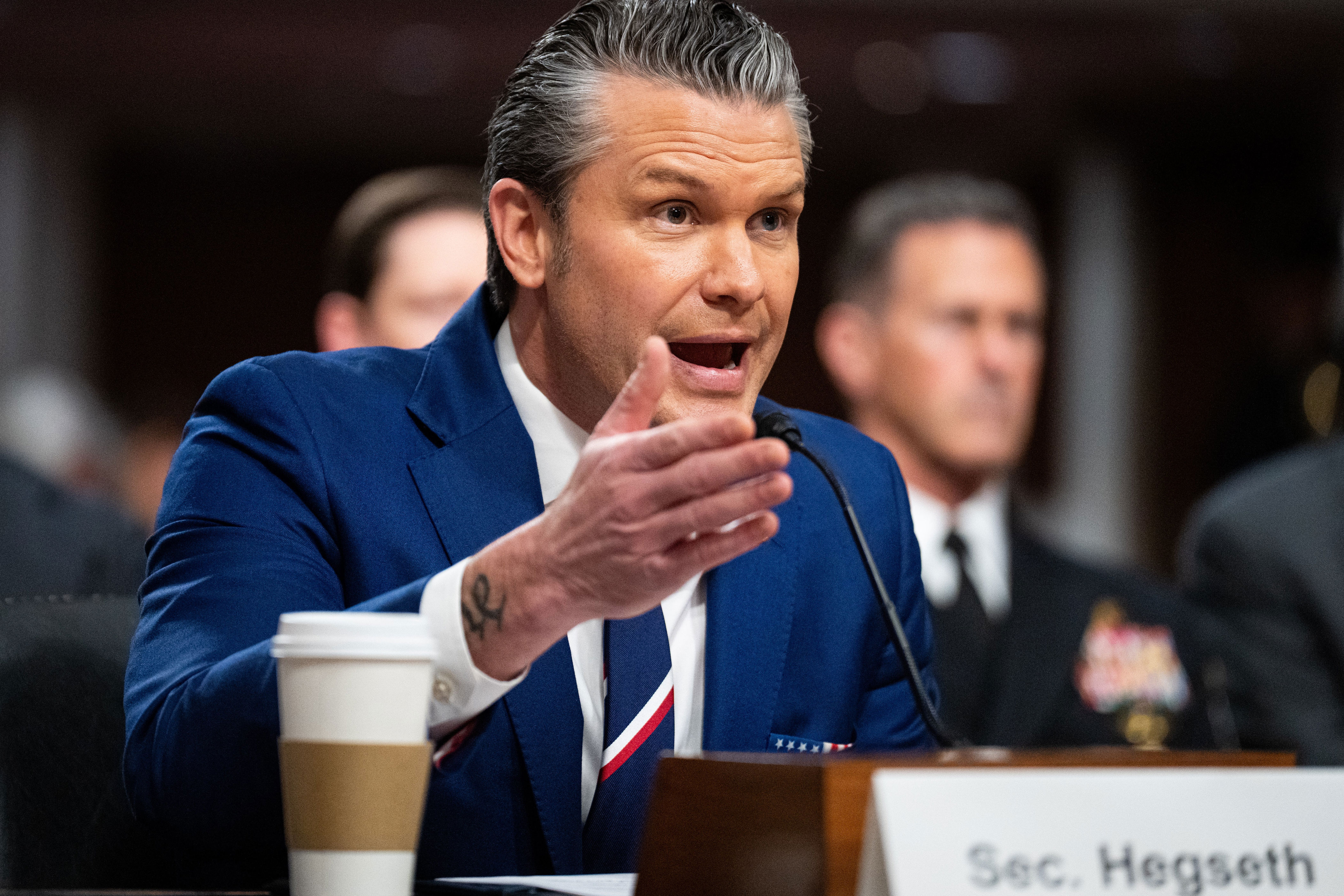 A person in a blue suit speaks passionately at a conference, gesturing with one hand. A coffee cup is on the table in front of them