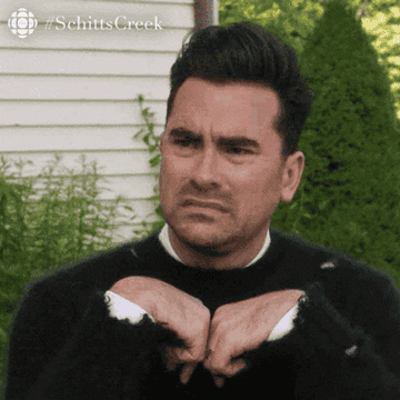 Person with a skeptical expression, hands near face, outdoors. Image labeled #SchittsCreek at the top, relating to a humorous or puzzling moment