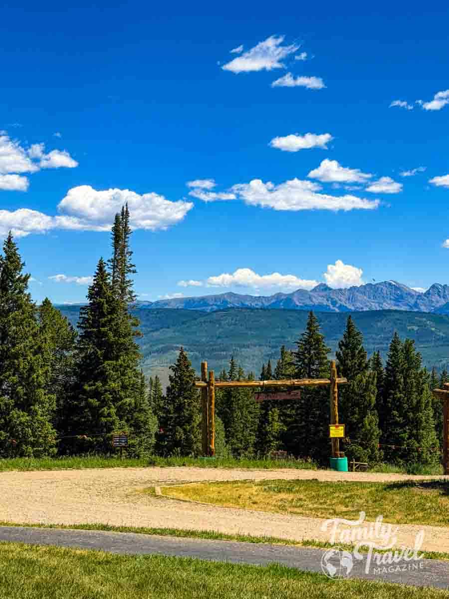 A wooden archway at Beaver Creek surrounded by pine trees and the Rocky Mountains under a blue sky.