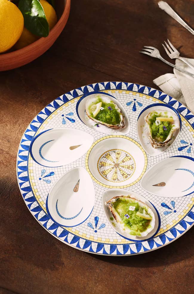 Decorative oyster plate holding oysters garnished with a green topping, set on a wooden table with a bowl of lemons and metal forks nearby