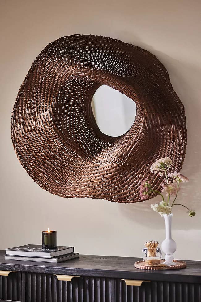 Woven wall mirror above a console table with a candle, books, and a vase with flowers, showcasing home decor essentials