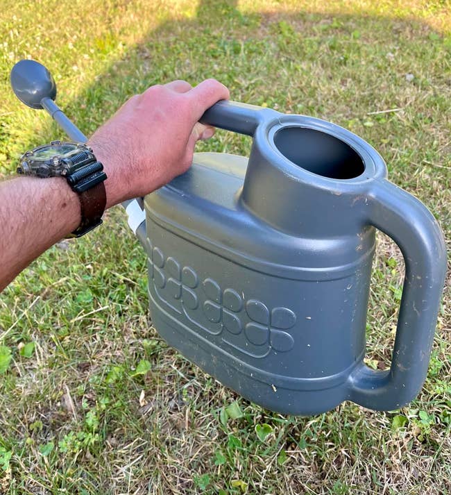 A person holding a gray watering can in a grassy area, wearing a watch on their wrist