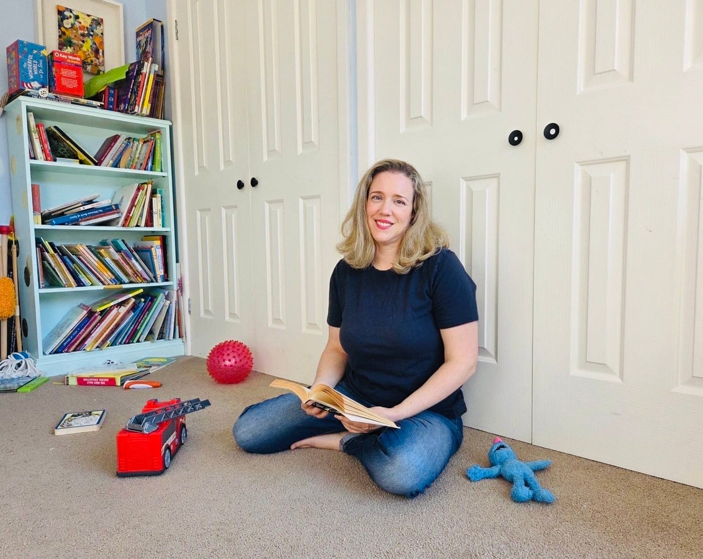 The author sits on the floor of a kid's room with an open book in her hand.