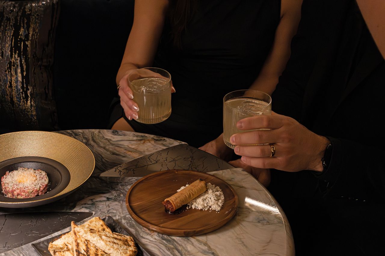 Two people holding glasses of drinks a table with appetizers and a dessert