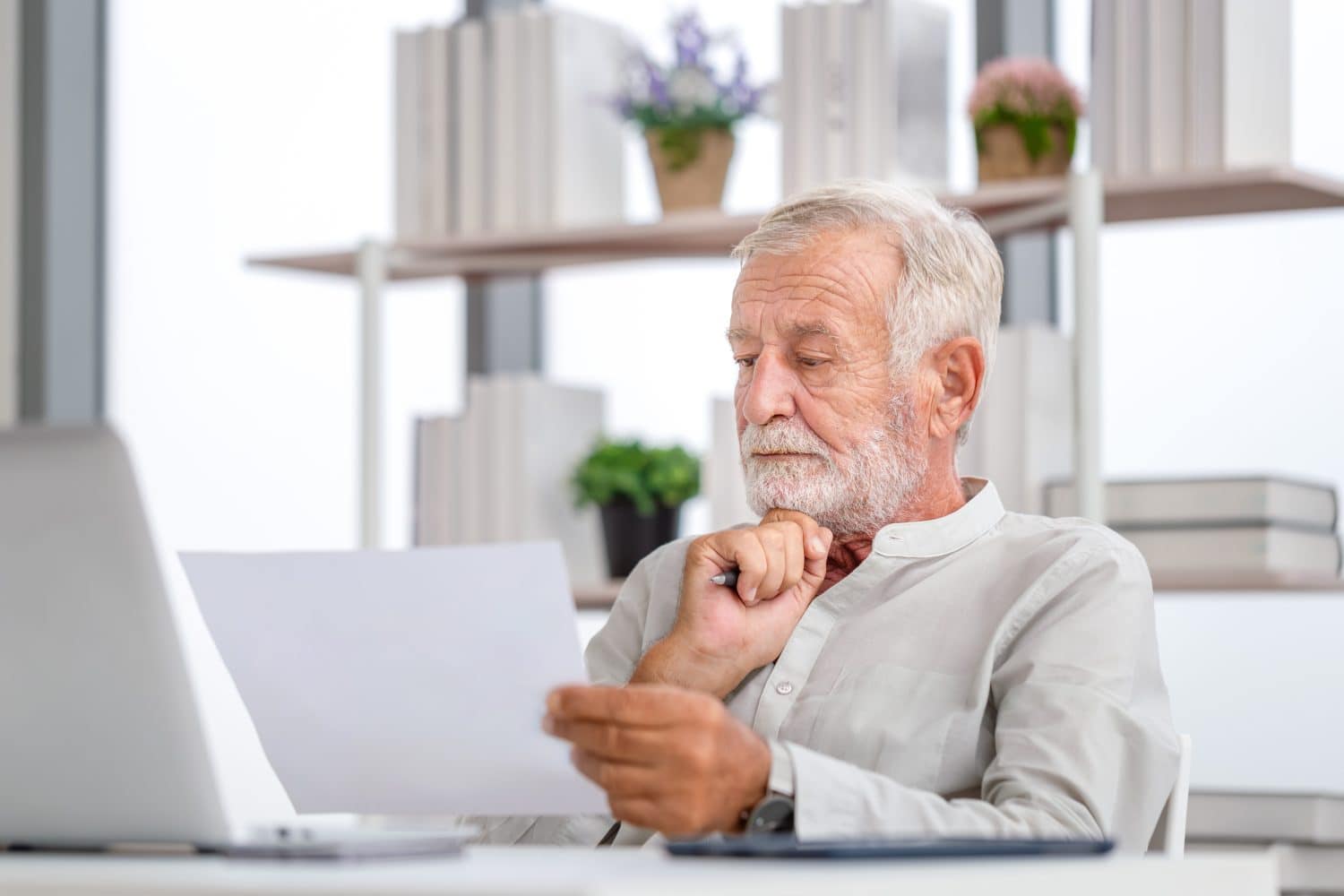 A close-up shot of an elderly man with gray hair and a white beard, wearing a light-colored collared shirt, intently looking down at a white paper he holds in his hands. He is holding a pen to his chin with his right hand, suggesting contemplation. A silver laptop is visible to his left, and a blurred background shows shelves with books and potted plants, indicating an office or home study setting.