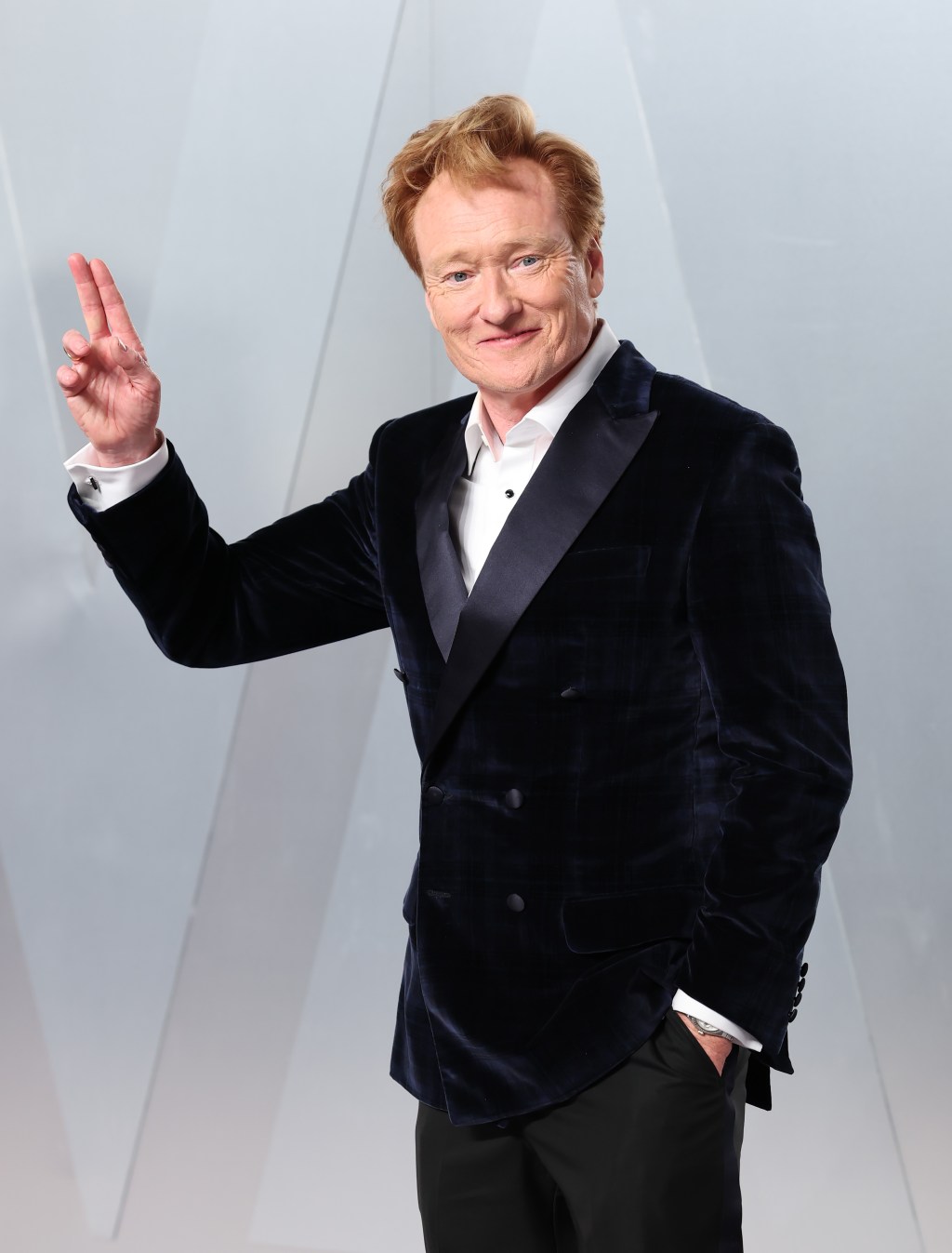 Conan O'Brien in a velvet tuxedo jacket smiles and holds up two fingers.