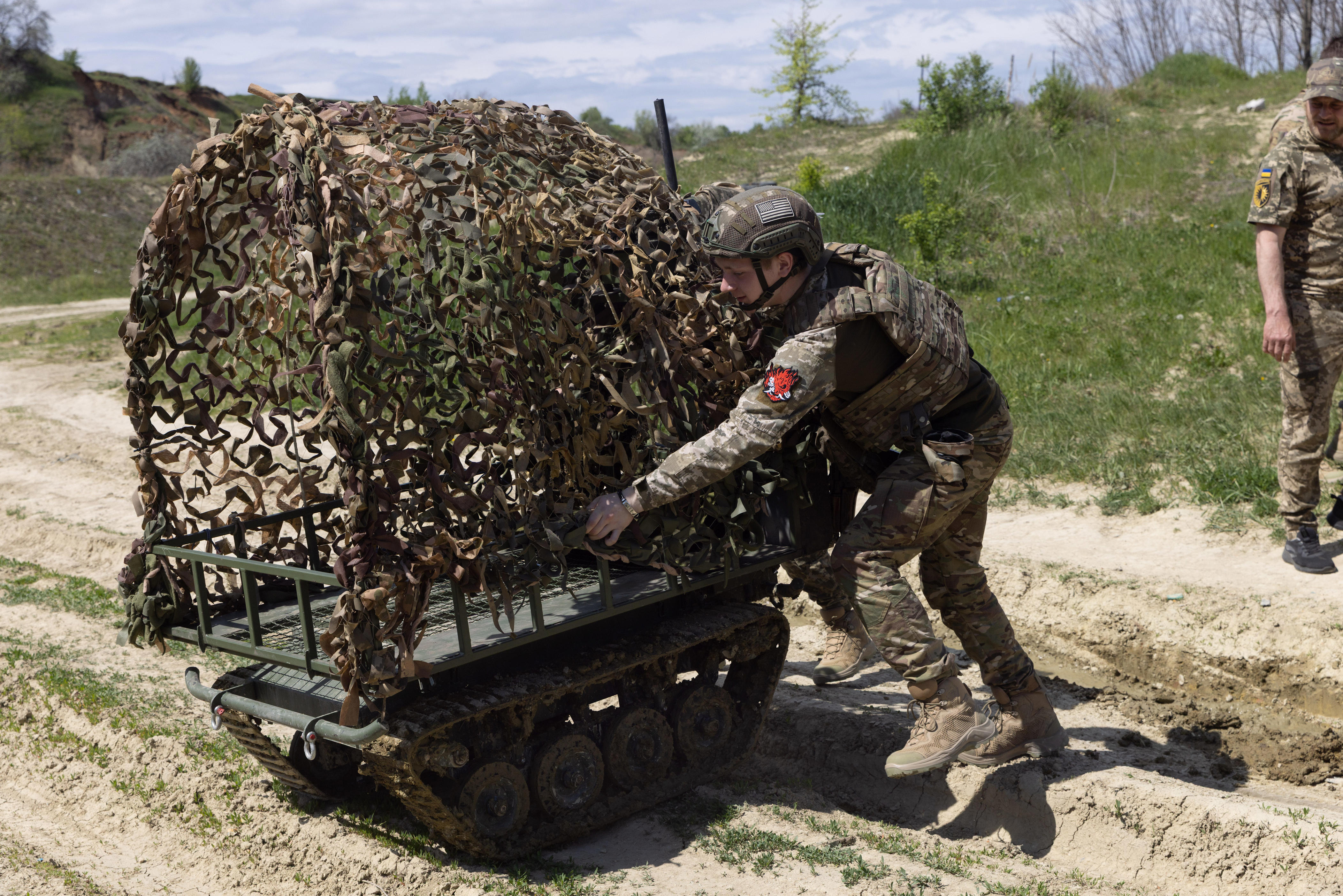 Uniformed personnel adjust camouflage netting on a small tracked ground vehicle along a dirt path.