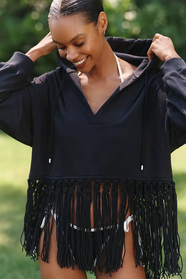 Model outdoors wearing a black fringe hoodie over a white bikini, adjusting the hood with a smile
