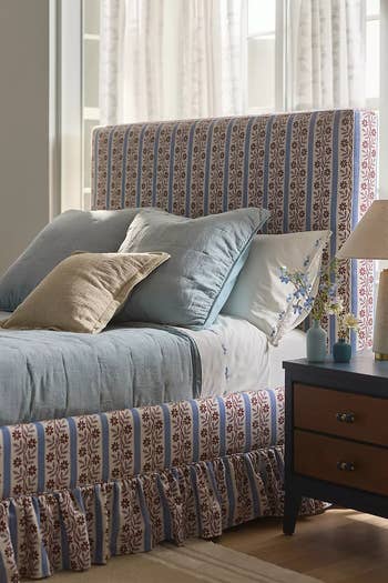 Patterned bed with pillows and a matching bed skirt in a cozy bedroom setting. Lamp and nightstand with decorative items beside the bed