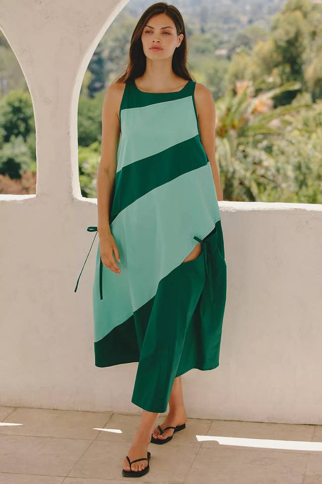 Model in a stylish, flowing green and turquoise dress with diagonal stripes, standing on a balcony with lush greenery in the background