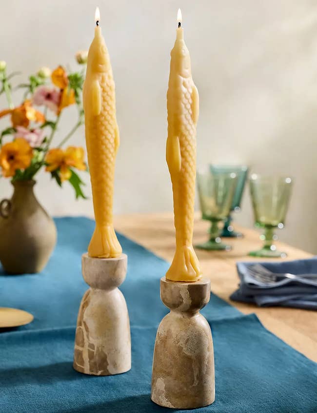 Fish-shaped candles on marble holders, set on a blue tablecloth with flowers and glasses in the background