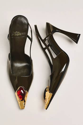High-heeled shoes with a glossy black finish, gold detailing, and a red jewel accent on the pointed toes, designed by Jeffrey Campbell