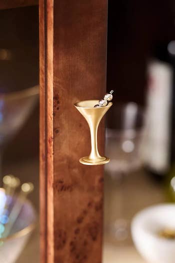 Gold champagne glass cabinet knob shown on a wooden surface. Blurred background includes a glass and dish, suggesting a refined home decor accessory