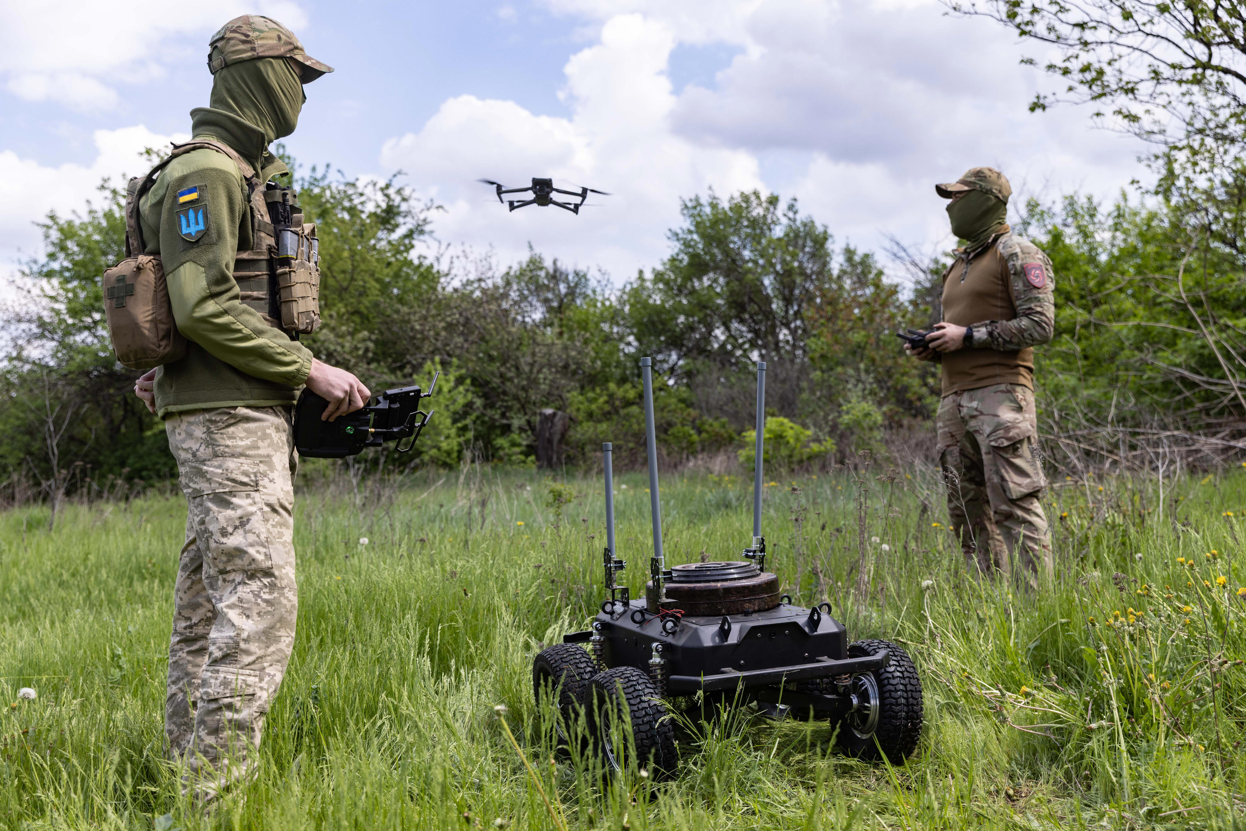 Two masked uniformed personnel operate an aerial drone and a ground robot in a grassy field.