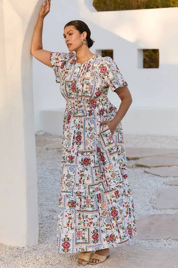 Person posing in a patterned, floral maxi dress with short flutter sleeves and pockets, standing outdoors against a minimalist background