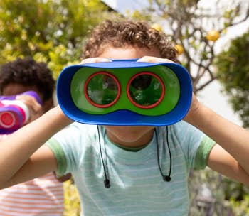 Child holding toy binoculars outdoors, looking directly at the camera. Another child is in the background, also holding binoculars