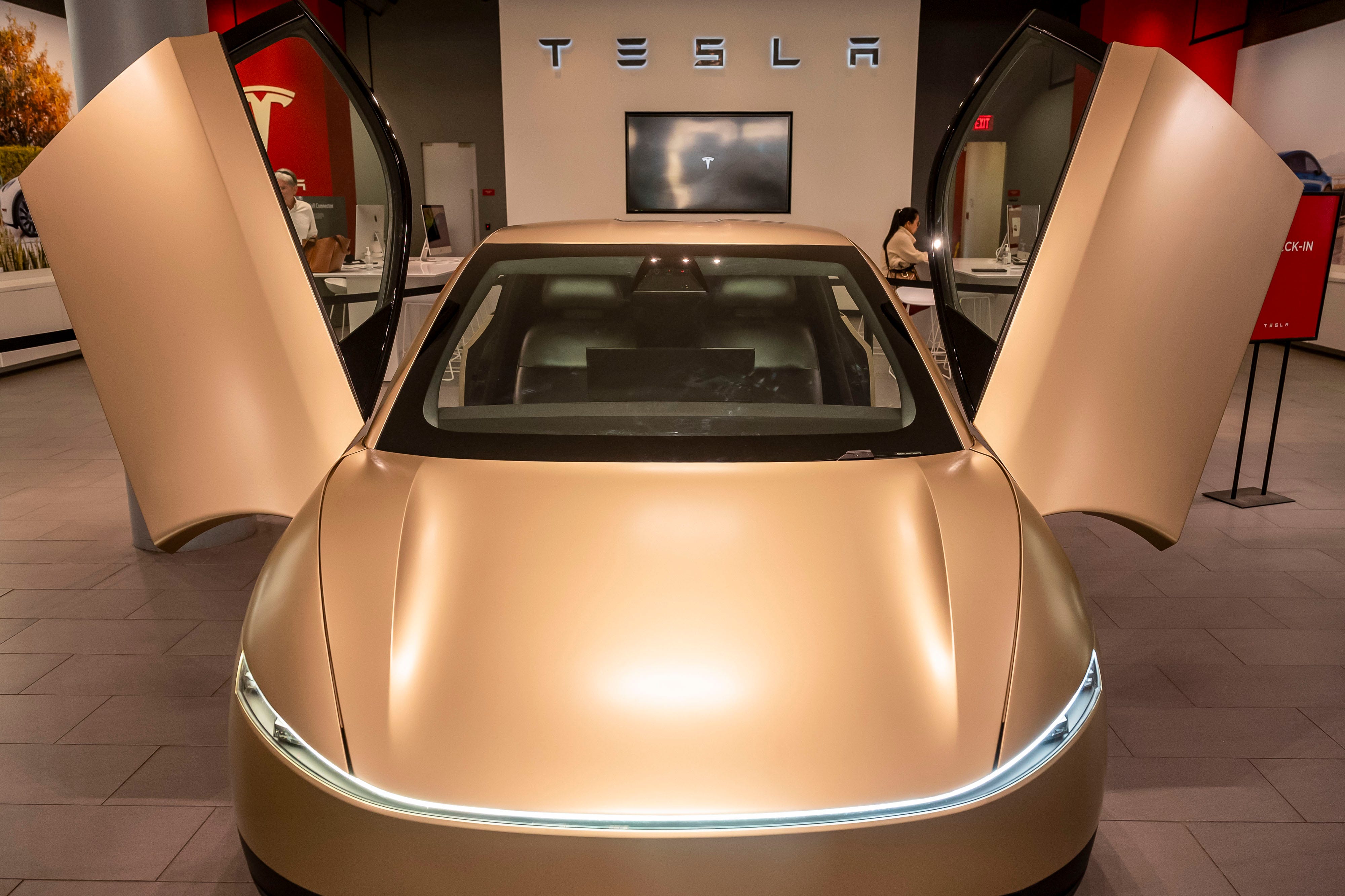 Gold Tesla robotaxi with both butterfly doors raised sits inside a showroom beneath T E S L A wall lettering.