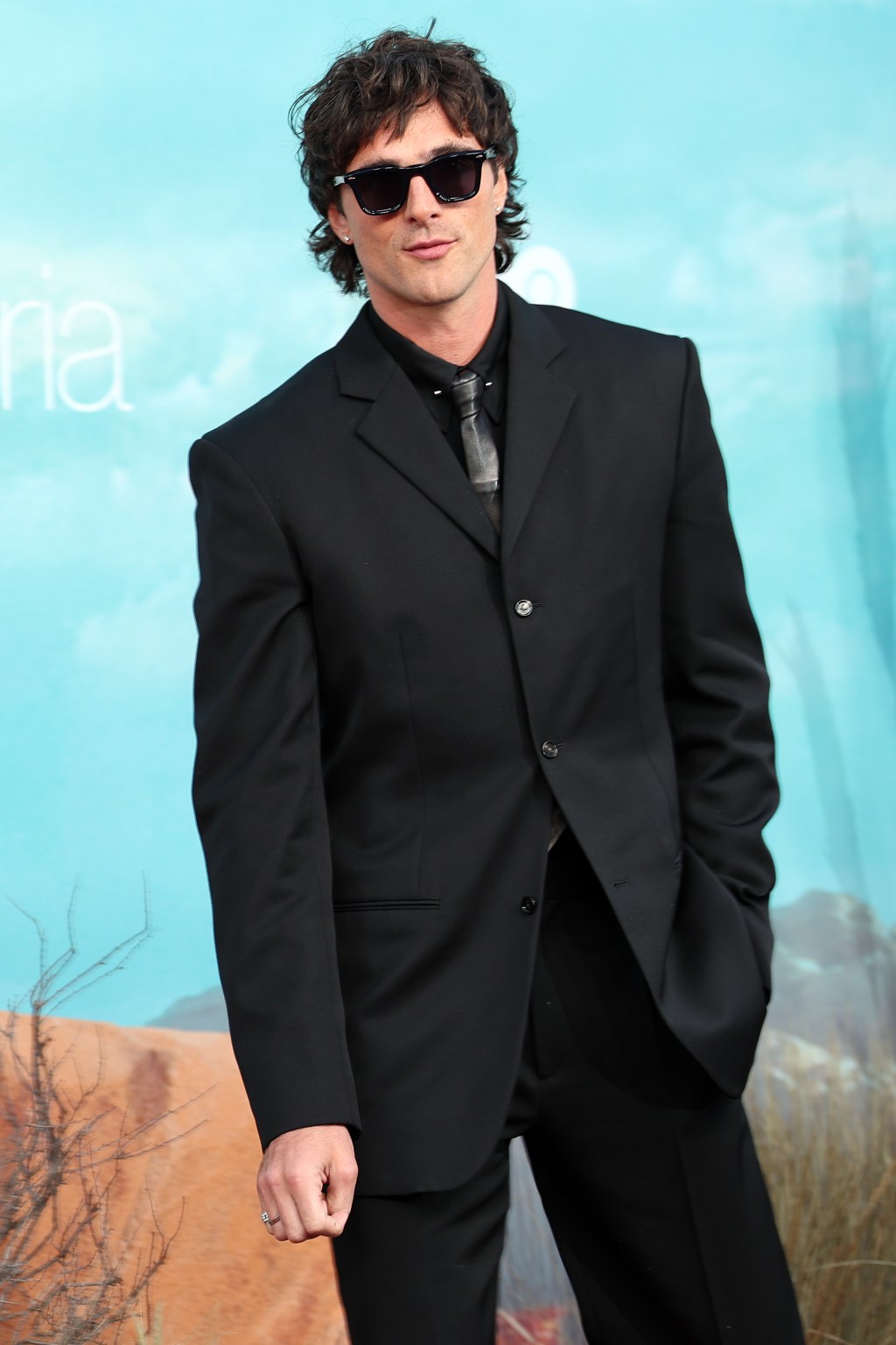 Jacob Elordi poses in a black suit, black shirt, and gray tie, wearing sunglasses.
