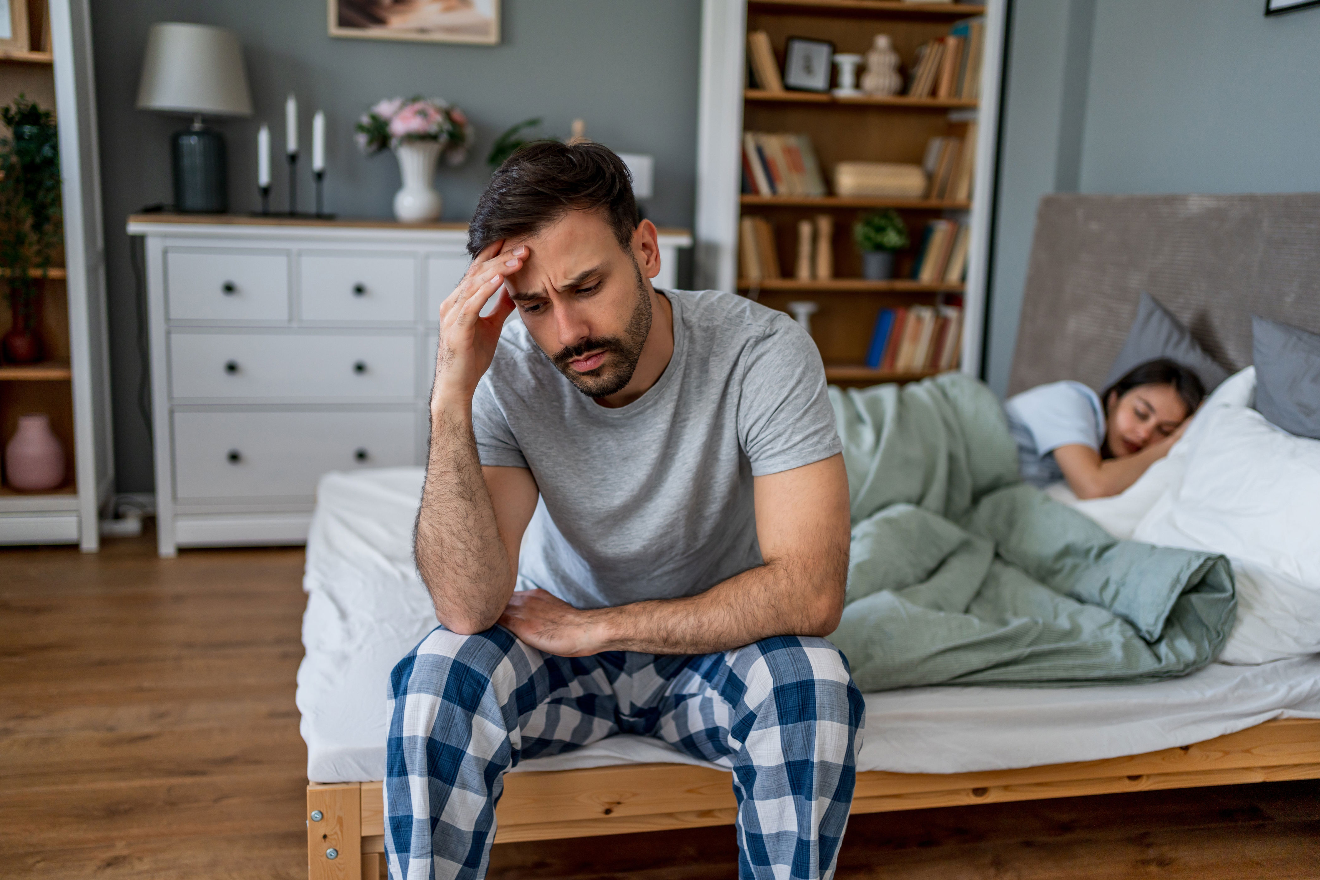 Man in pajamas sits on bed with a pensive expression, while a woman sleeps next to him, suggesting a possible personal or relationship issue