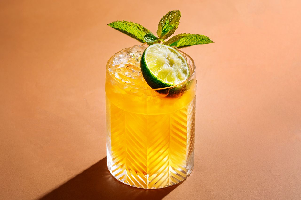 A Mai Tai cocktail with half of a lime and a sprig of mint on a peach colored background.