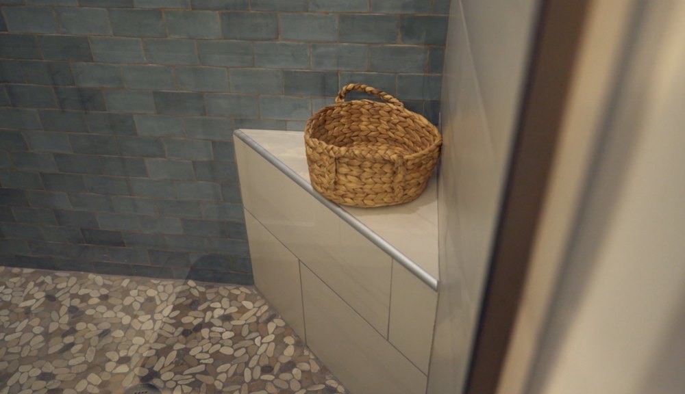 The primary suite’s full-sized bathroom has a pebble tile shower pan, various blue-toned subway tiles on the wall, and oversized white subway tiles covering a corner seat.