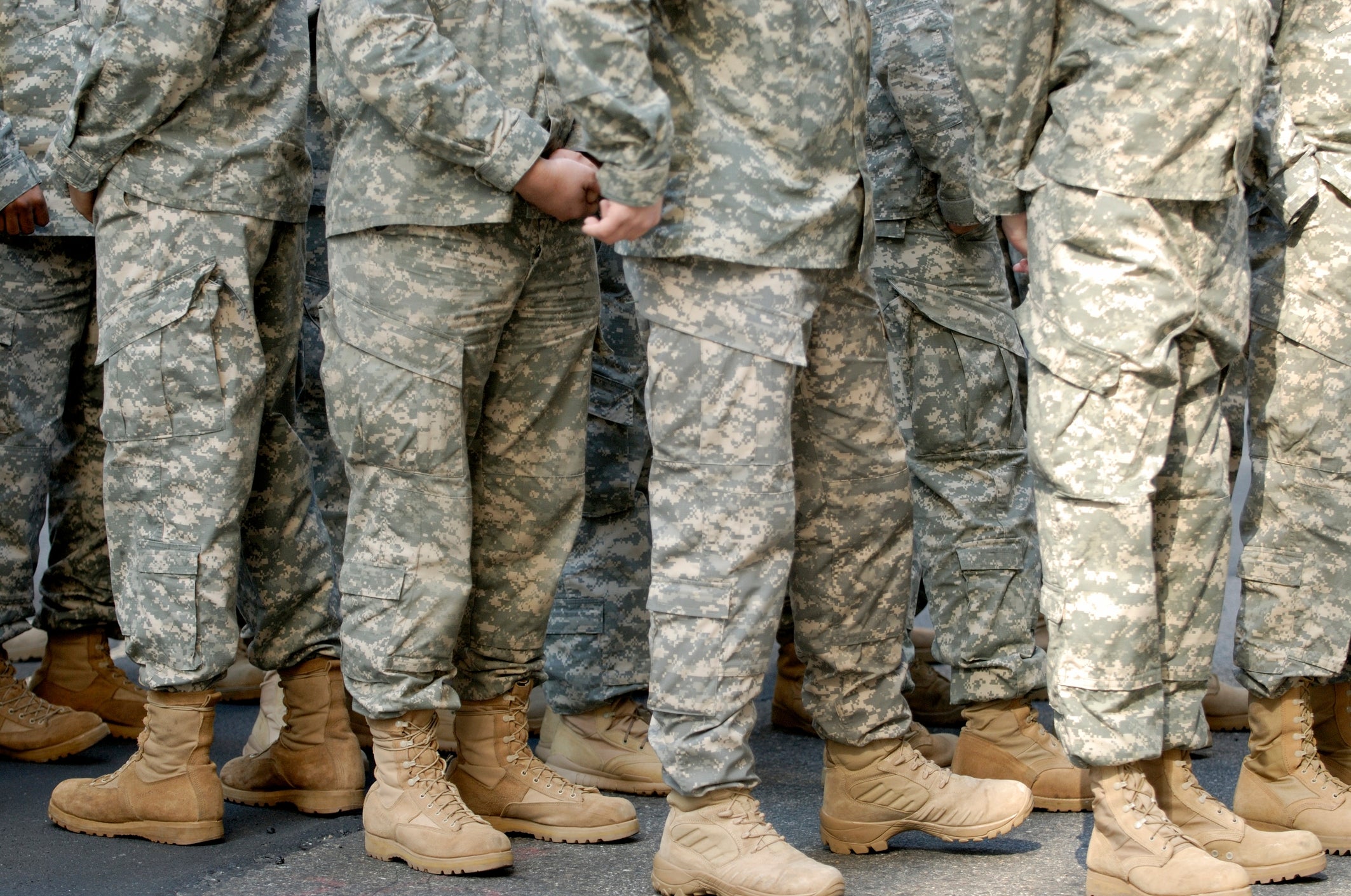 People in military uniforms stand in formation, wearing camouflaged fatigues and tan boots