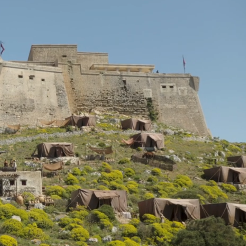 Stone fortress on a hill with scattered tents and structures at its base, under a clear sky