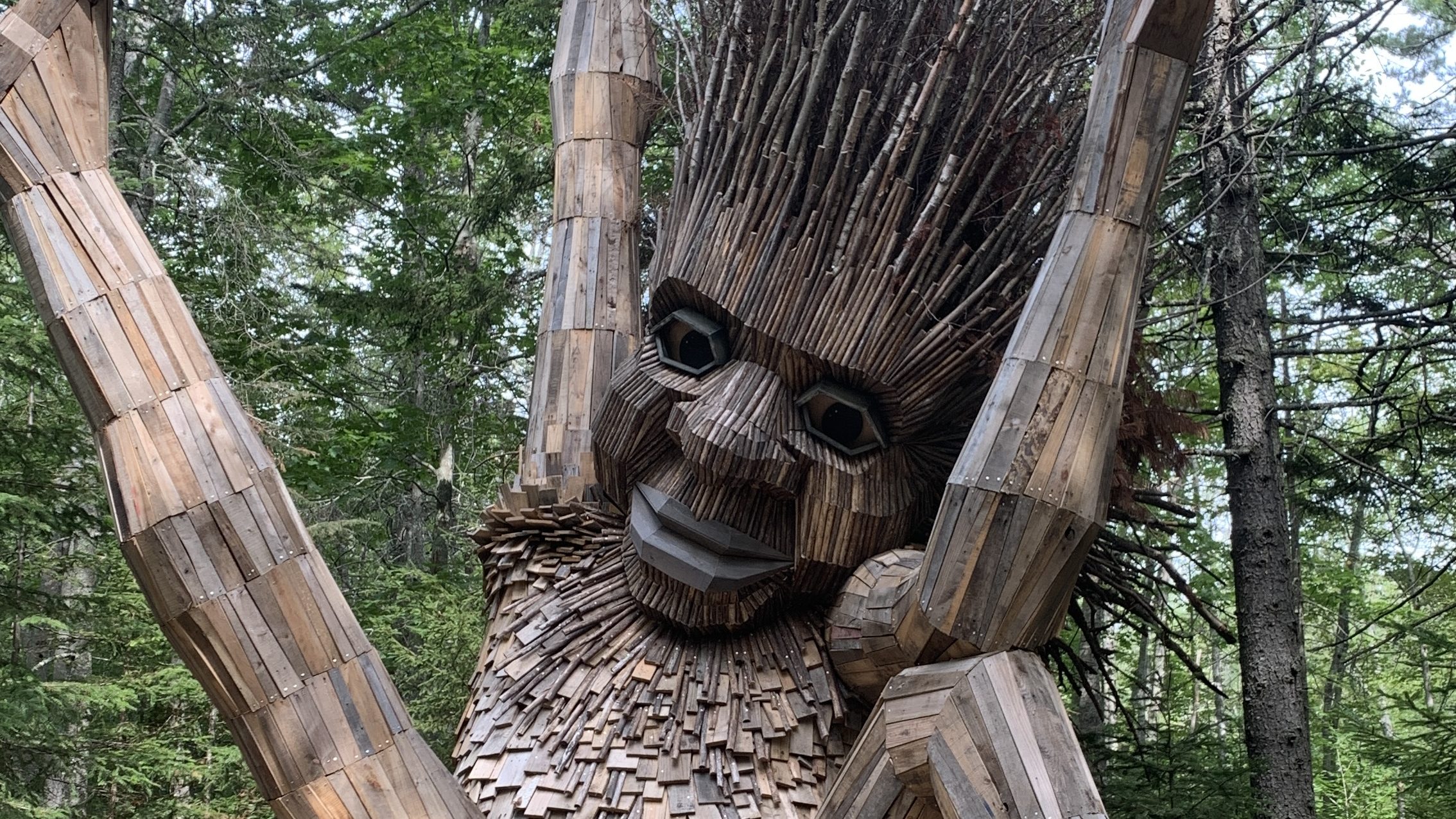 A giant wooden troll sitting in the forest with both arms lifted and even a leg lifted in an exuberant expression of joy.