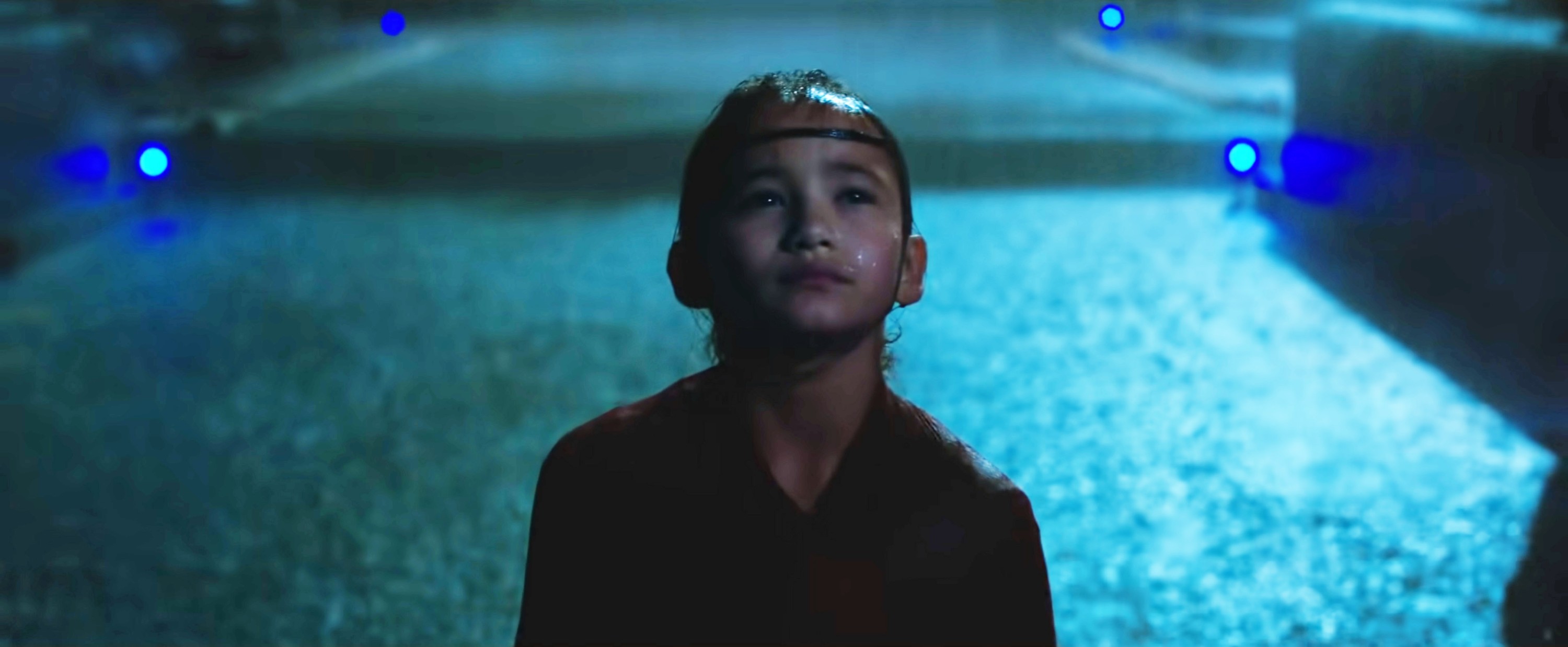 Kaylee Hottle as Jia, a young girl with a headband, standing in the rain and looking up, with blue lights in the background.