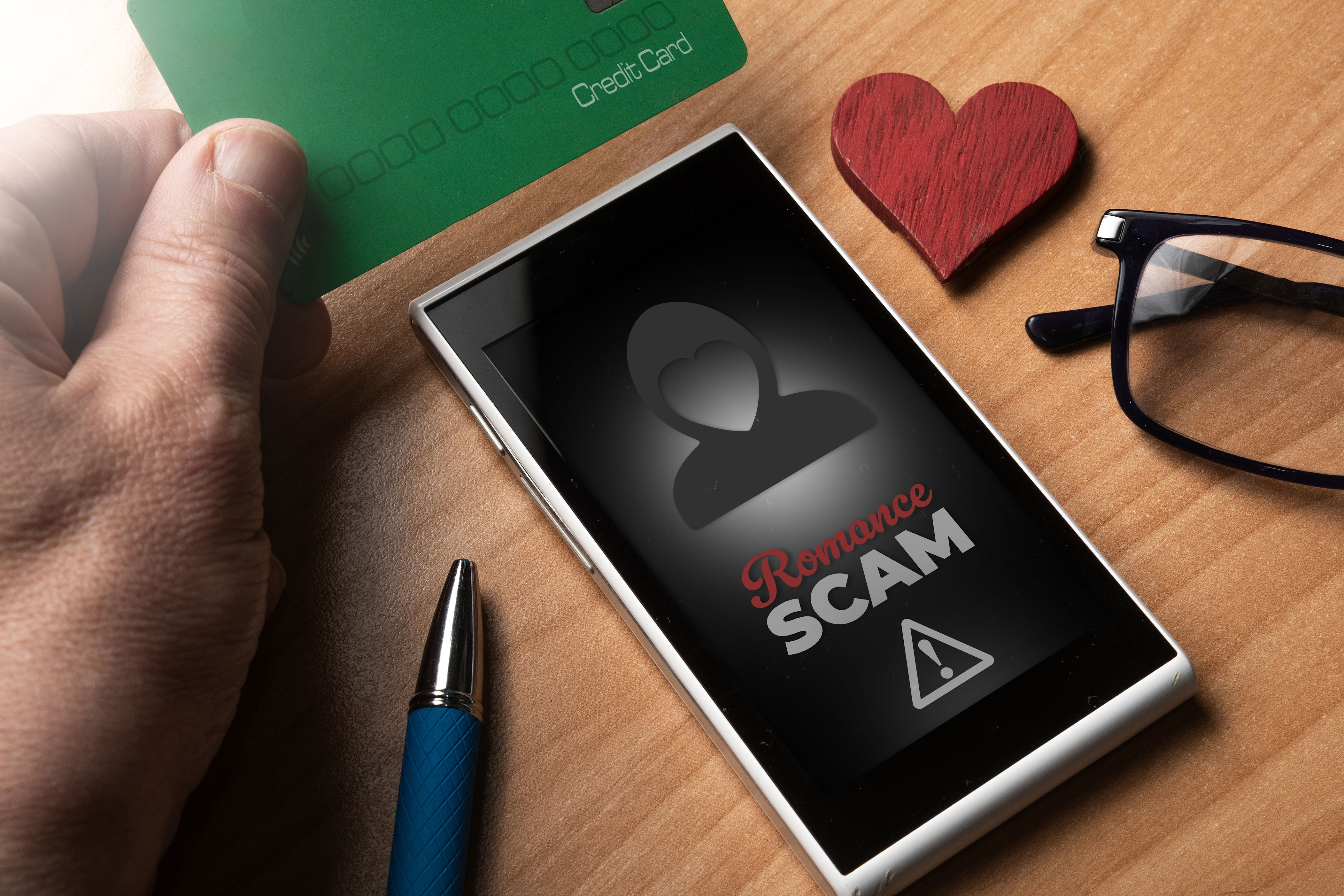 Smartphone displaying "Romance Scam" warning message; hand holds credit card nearby. Glasses, pen, and heart-shaped object on table