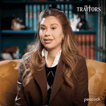 A person sits on a couch in a library setting, expressing surprise. Text overlay reads: "WHAT A FLIP!" "The Traitors" logo is visible