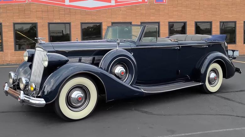 A 1937 Packard Super Eight Convertible Sedan finished in dark blue, parked in an empty lot outside a brick building.