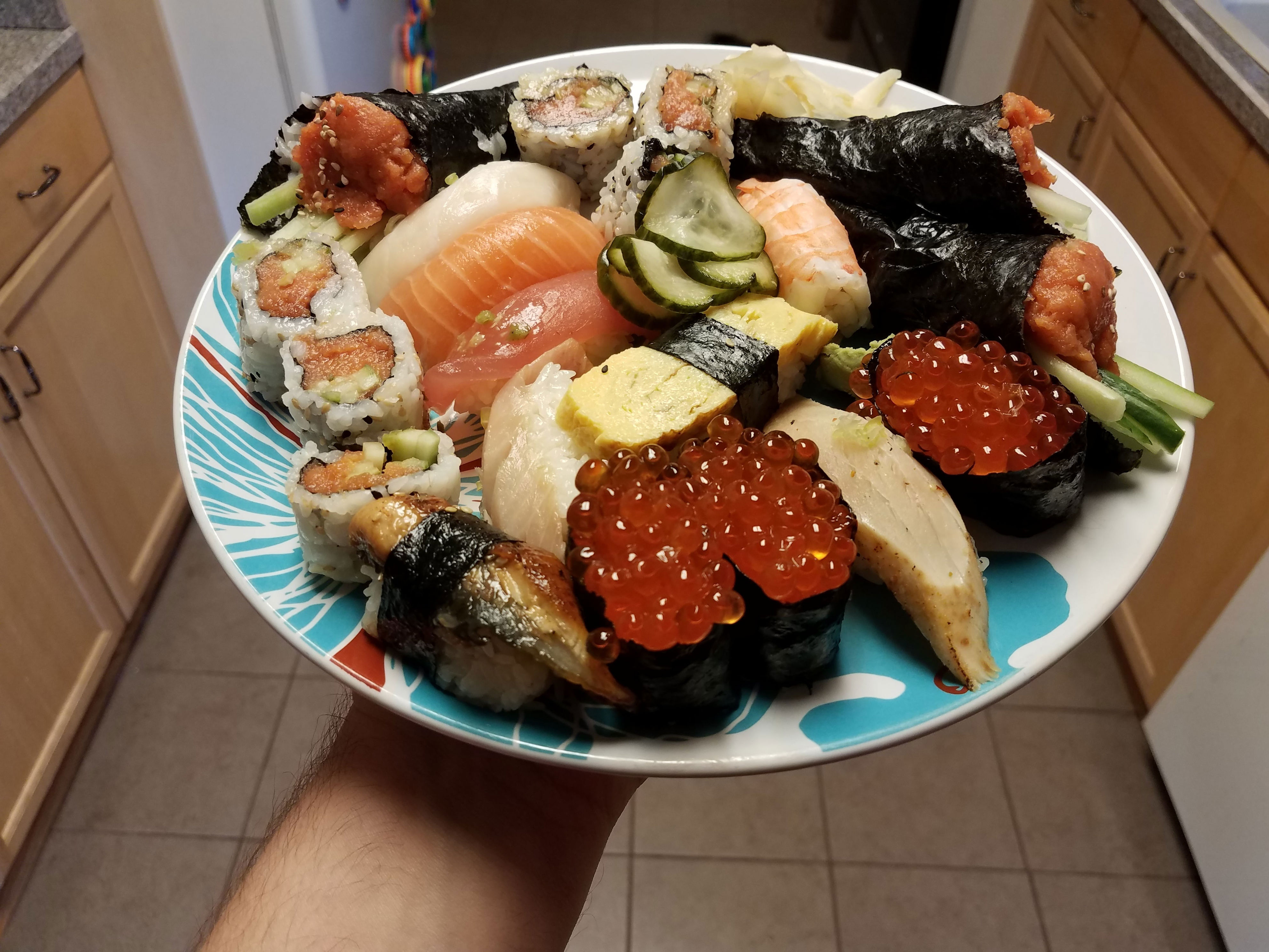 A plate of assorted sushi held over a kitchen floor, featuring nigiri, rolls, and garnishes