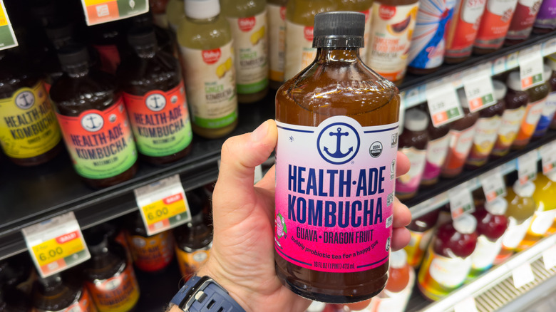 A hand holding up a bottle of Health-Ade kombucha in a store with other kombuchas in the background