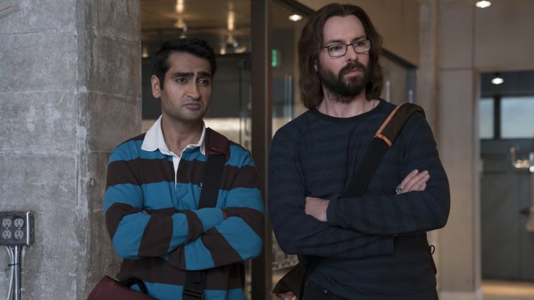 Kumail Nanjiani and Martin Starr as Dinesh and Gilfoyle standing side-by-side with arms crossed and skeptical expressions in a modern office in a scene from "Silicon Valley."