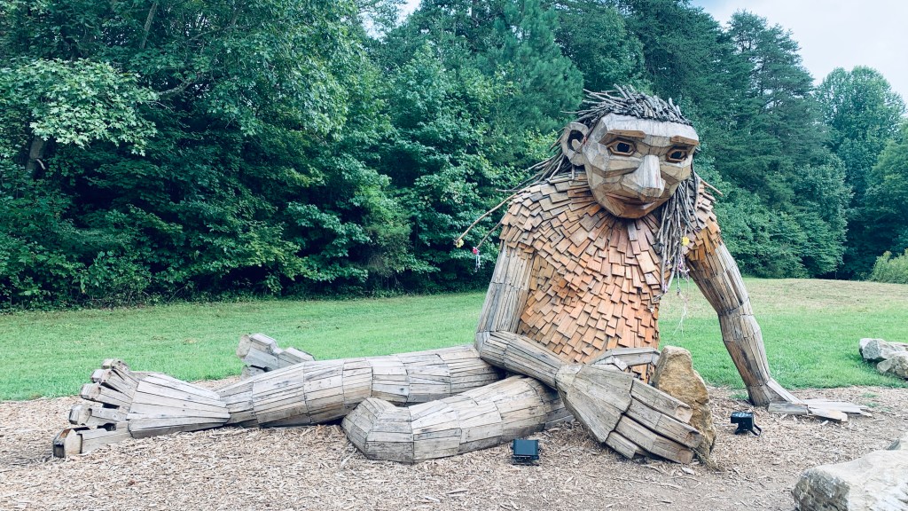 A wooden troll statue lounges along the ground while playing with rocks.