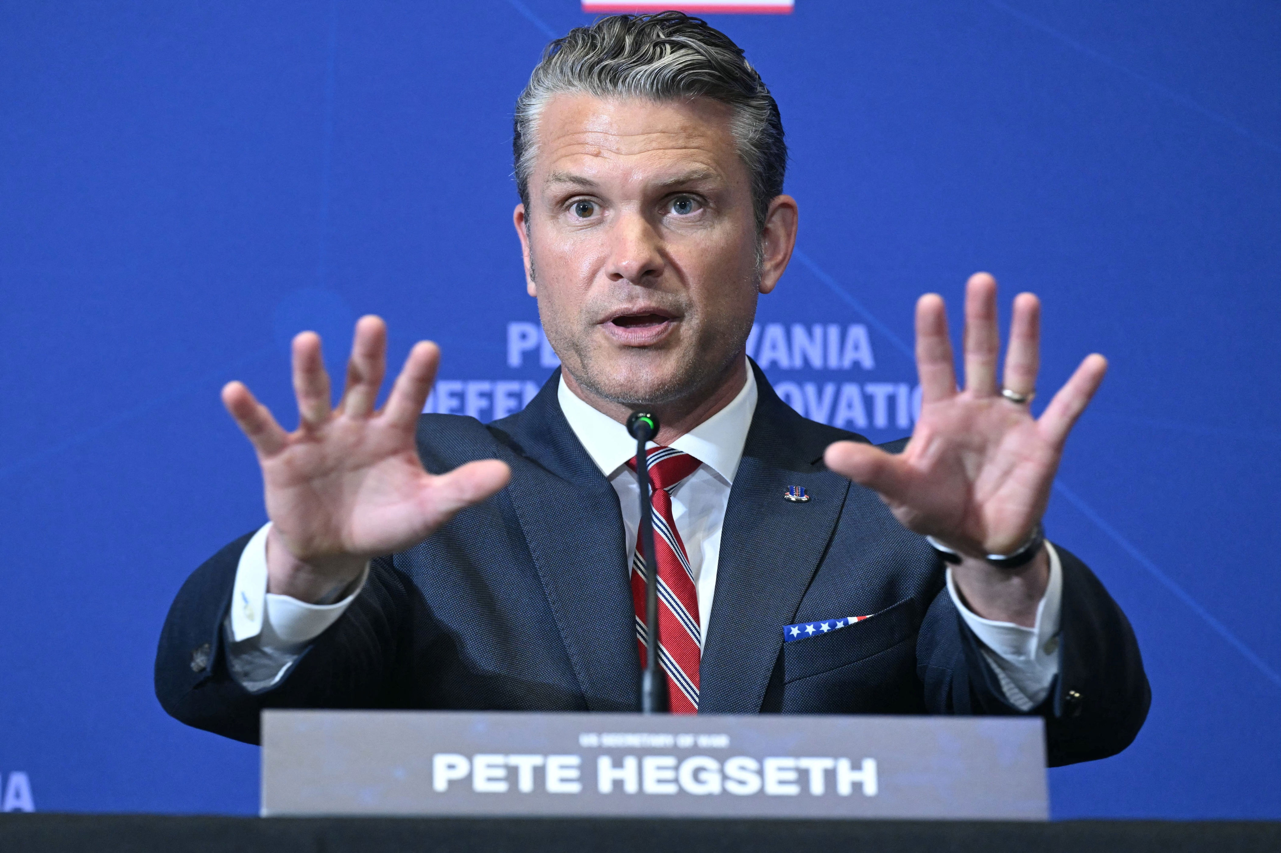 A man in a suit and striped tie speaks at a panel event, gesturing with both hands. A nameplate reads "Pete Hegseth."