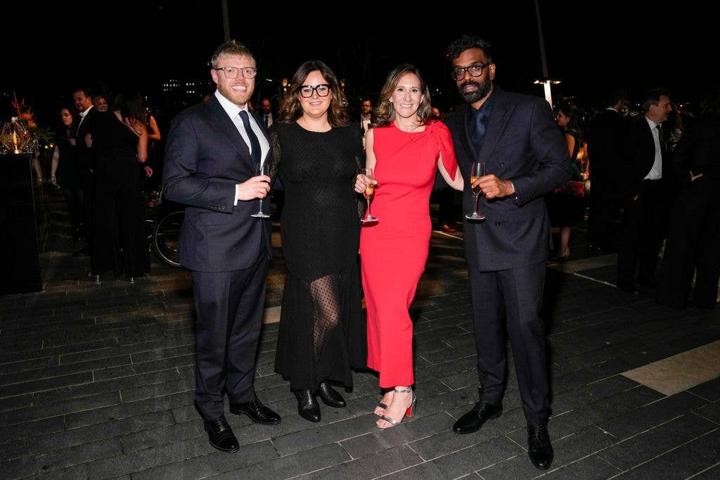 Four people in formal wear, holding wine glasses, at an outdoor event. The man on the right wears glasses.