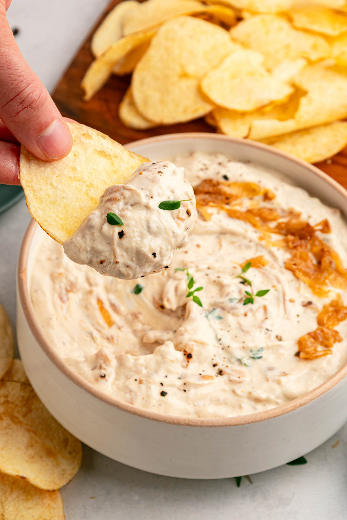 A hand dips a potato chip into a bowl of creamy onion dip, with more chips and fresh herbs visible in the background.