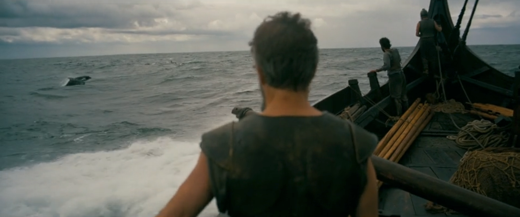 A person stands on a wooden ship's deck looking out at the ocean, with a whale breaching in the distance