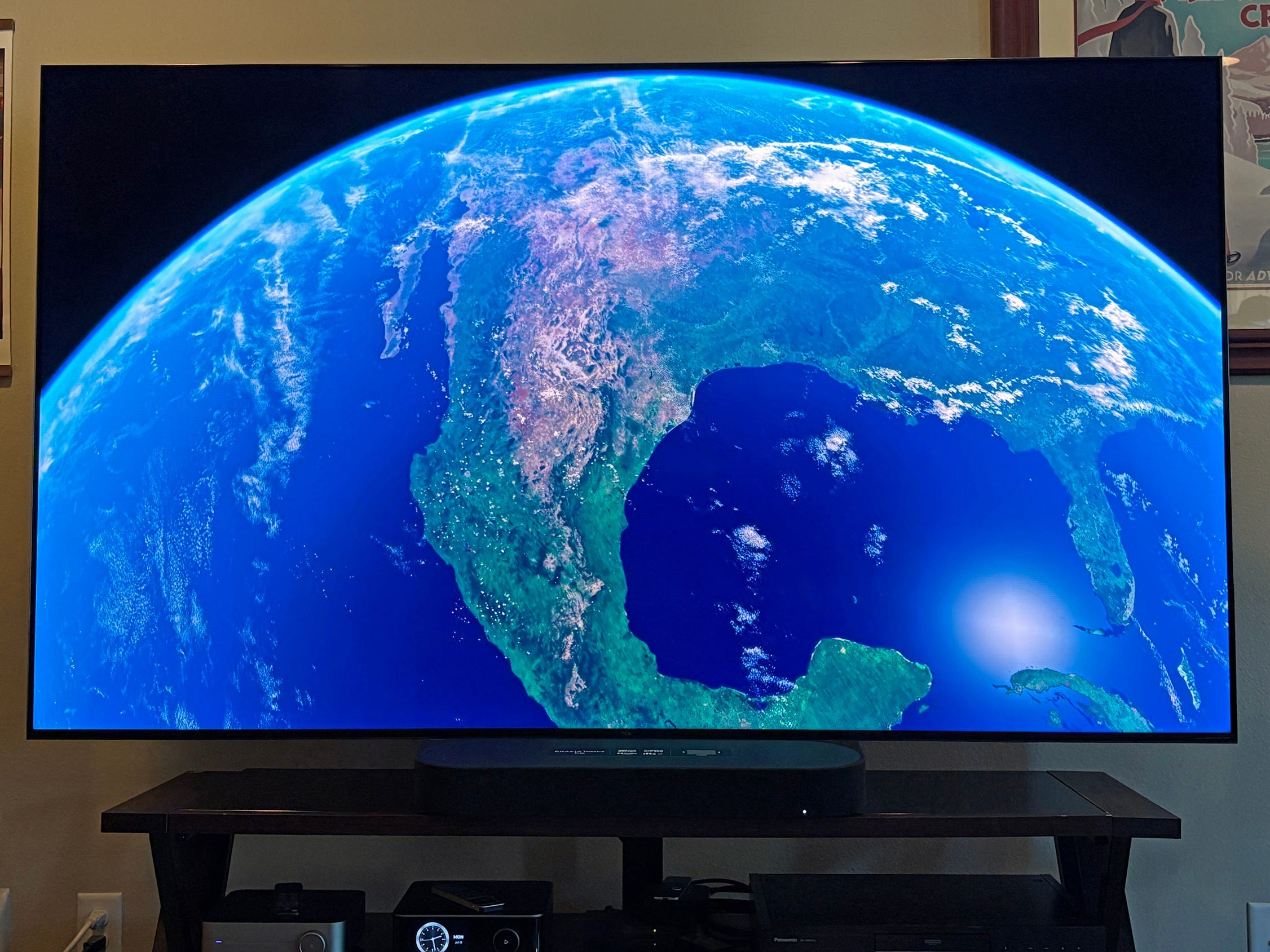 A TCL QM7L TV displays a satellite view of Earth in our reviewer's living room entertainment setup with speakers and framed wall art.