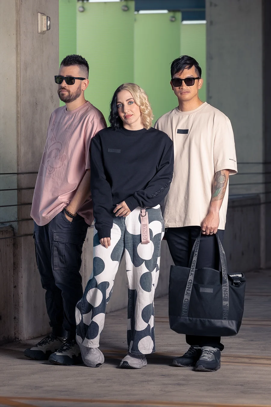 Three people stand together in casual attire, wearing sunglasses, in an urban setting with a green background.