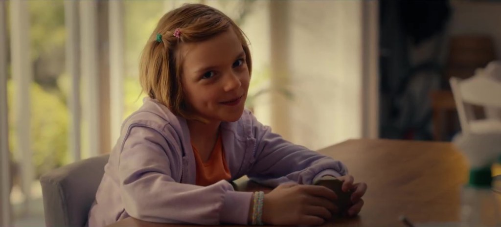 A young girl with blonde hair in pigtails smiles while sitting at a table.