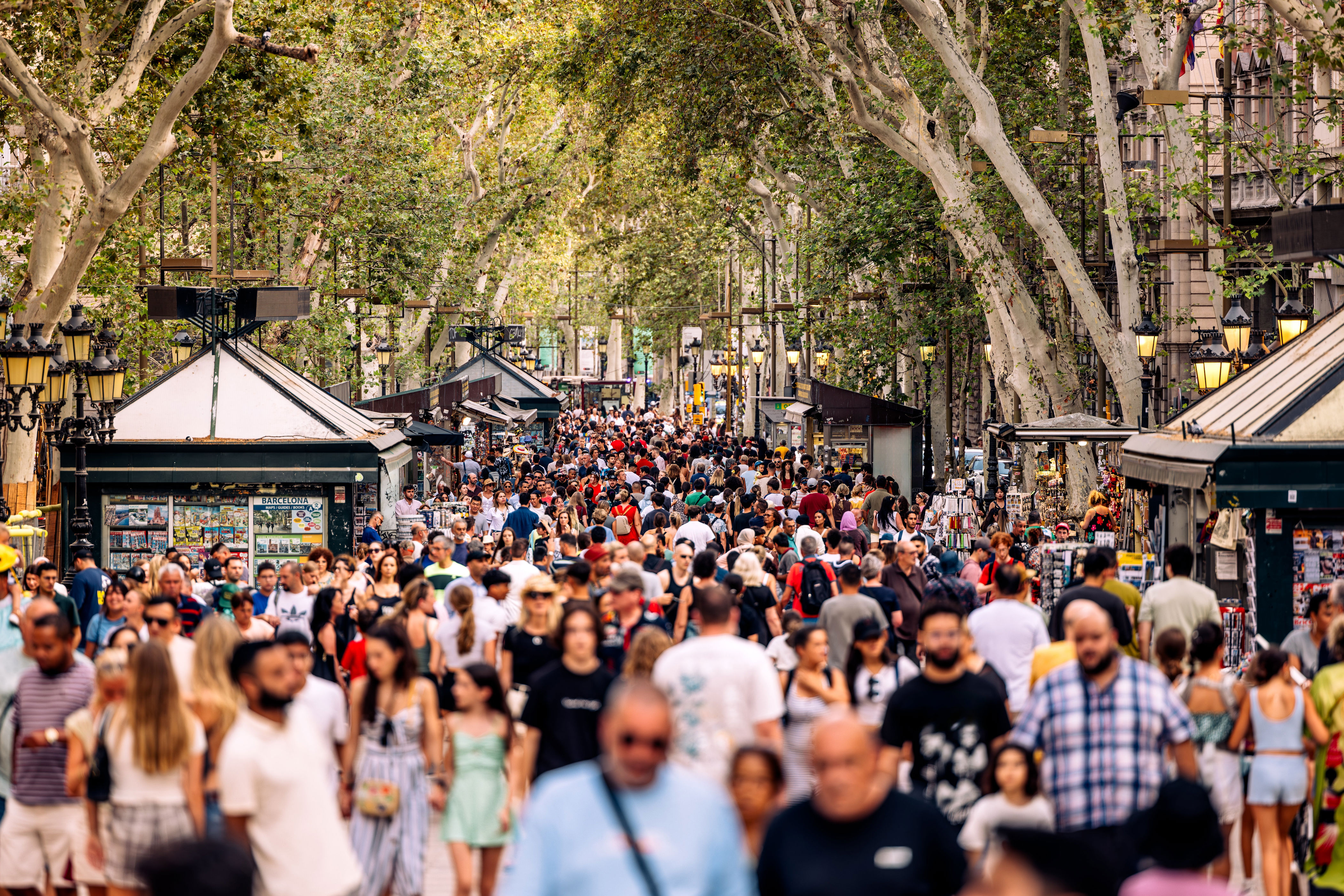 Crowded street in Barcelona lined with kiosks and trees, bustling with tourists and locals enjoying a lively atmosphere