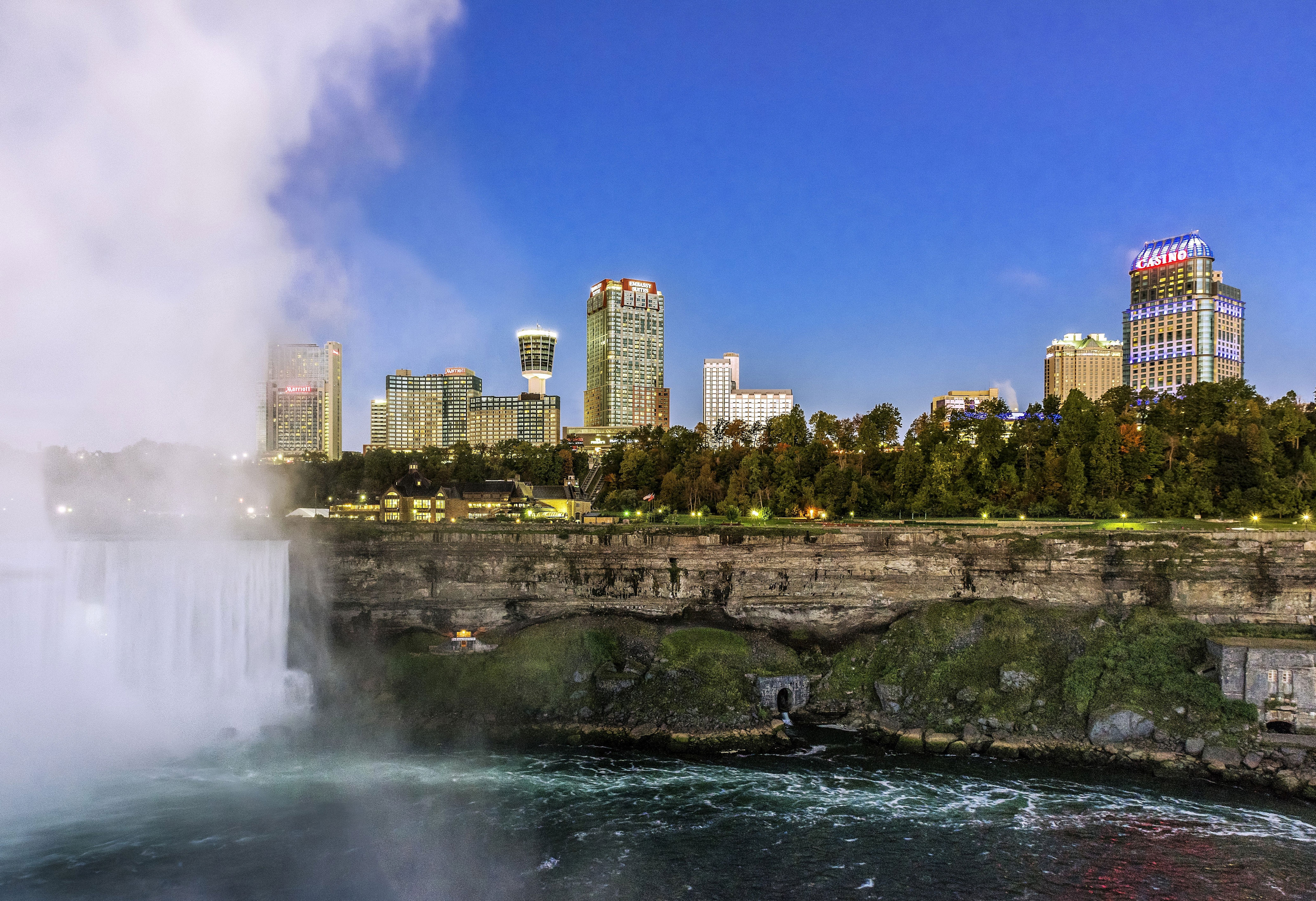 Cityscape view of buildings and skyline near a waterfall, with lush greenery and mist in the foreground