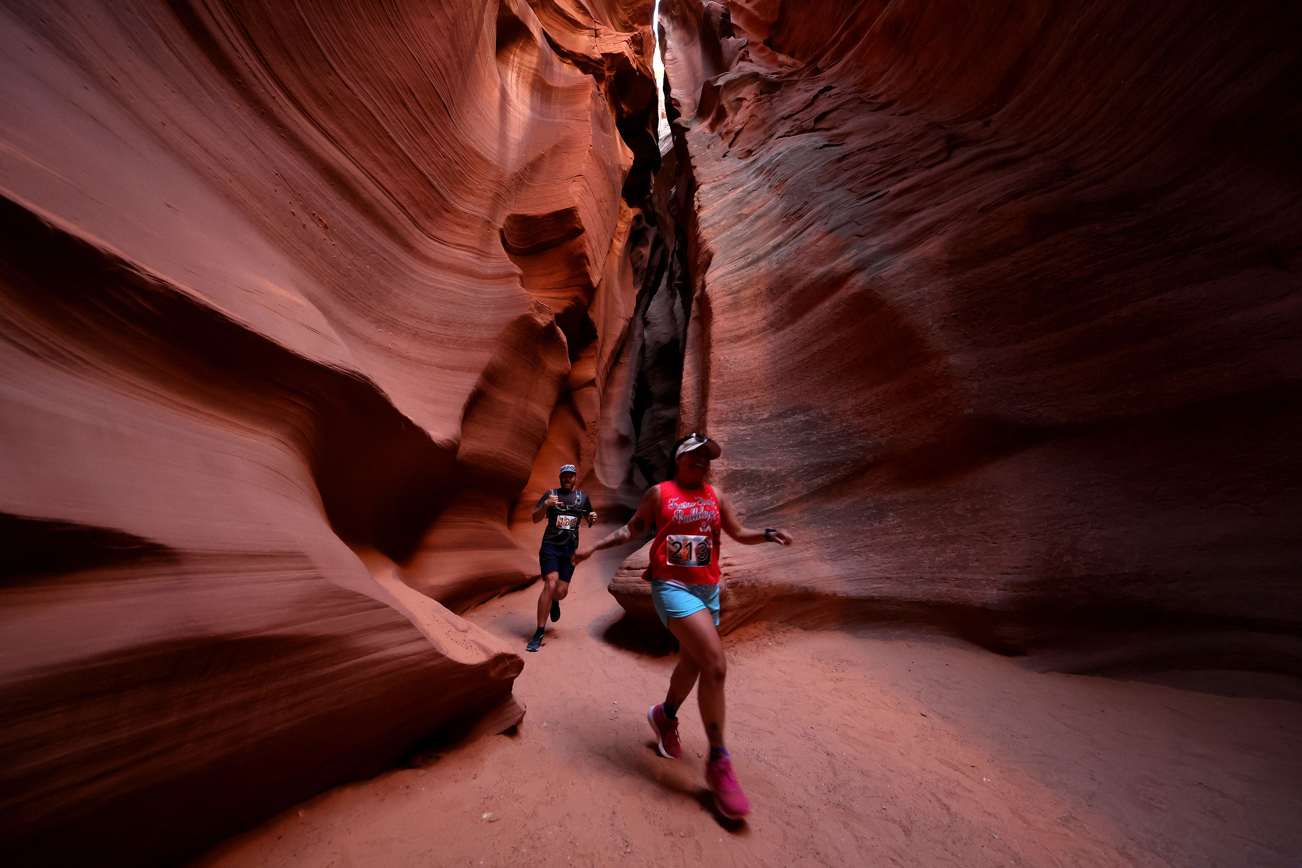 Two people run through narrow, winding sandstone canyons during a race, surrounded by tall rock walls