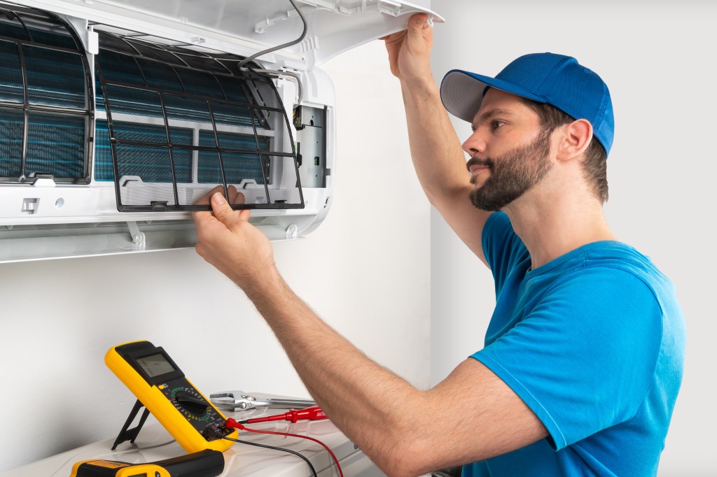 Installation service fix repair maintenance of an air conditioner indoor unit, by cryogenist technican worker checking the air filter in blue shirt baseball cap