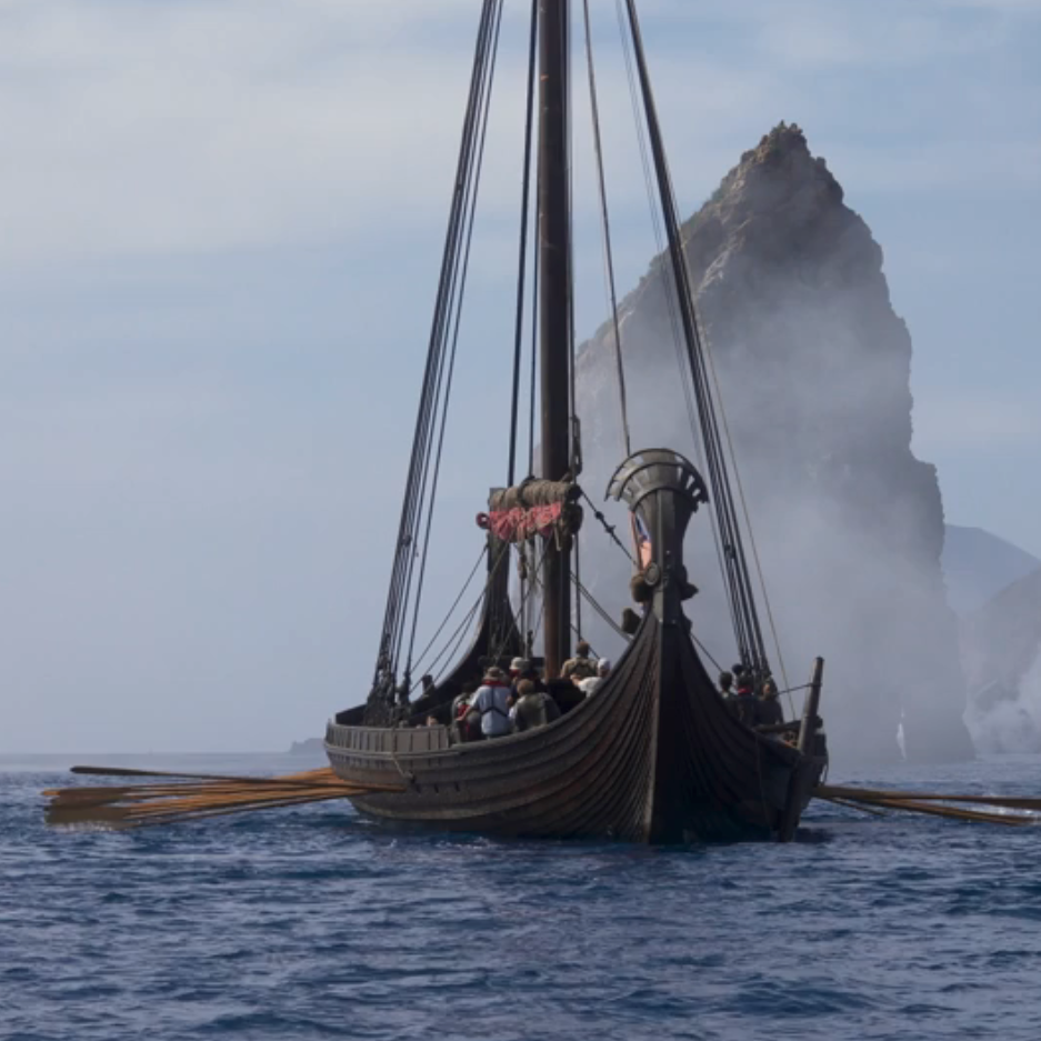 A Viking longship with a dragon head prow sails through misty waters near a jagged rock formation, with a small modern boat nearby