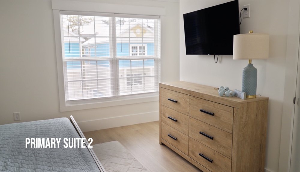 A second bedroom houses a bed with a blue blanket that sits opposite a wood-toned dresser and flat screen TV mounted on more creamy white walls.